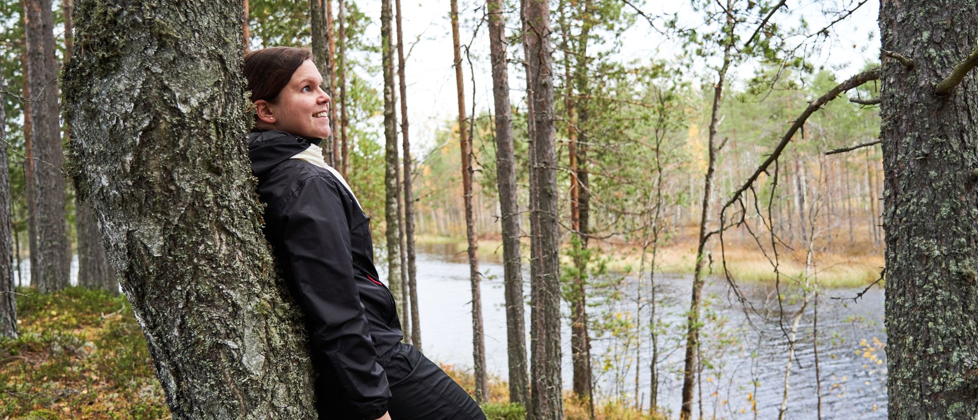 a hiker leaning against a tree, forest and lake in the background